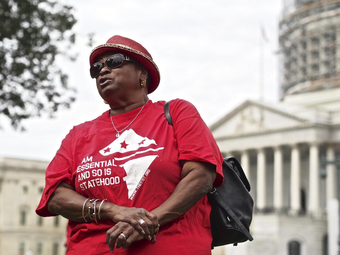 A woman stands in front of the U.S. Capitol wearing a shrit that says " I Am Essential and so Is Statehood "