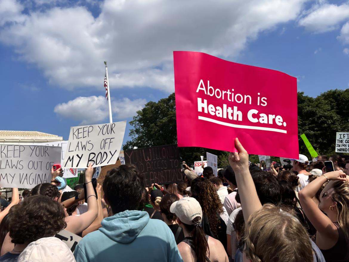 A crowd of people outside at an abortion rights rally. Someone is holding a sign that says Abortion is Health Care. Someone else holds a sign saying Keep Your Laws Off My Body. 
