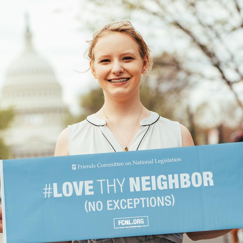 Advocate smiles in front of U.S. Capitol, holds sign that says: "Love They Neighbor (No Exceptions)"