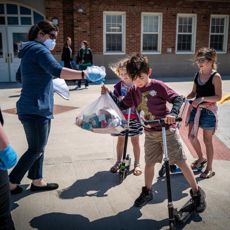 Des Moines Public Schools launched a huge increase in the number of free meal sites for students during the COVID-19 closure, going from 22 to 50 locations around the city. Here students on scooters receive a bag of food from volunteers.