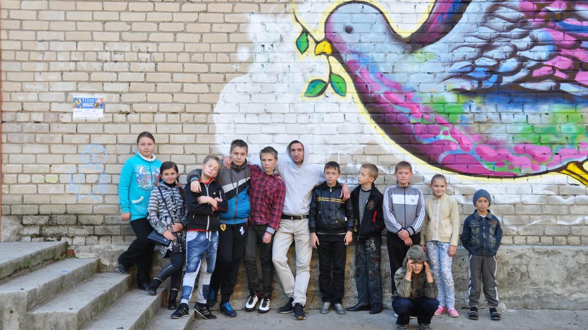 Kids in New Donbass, Ukraine stand in front of a mural depicting a peace dove